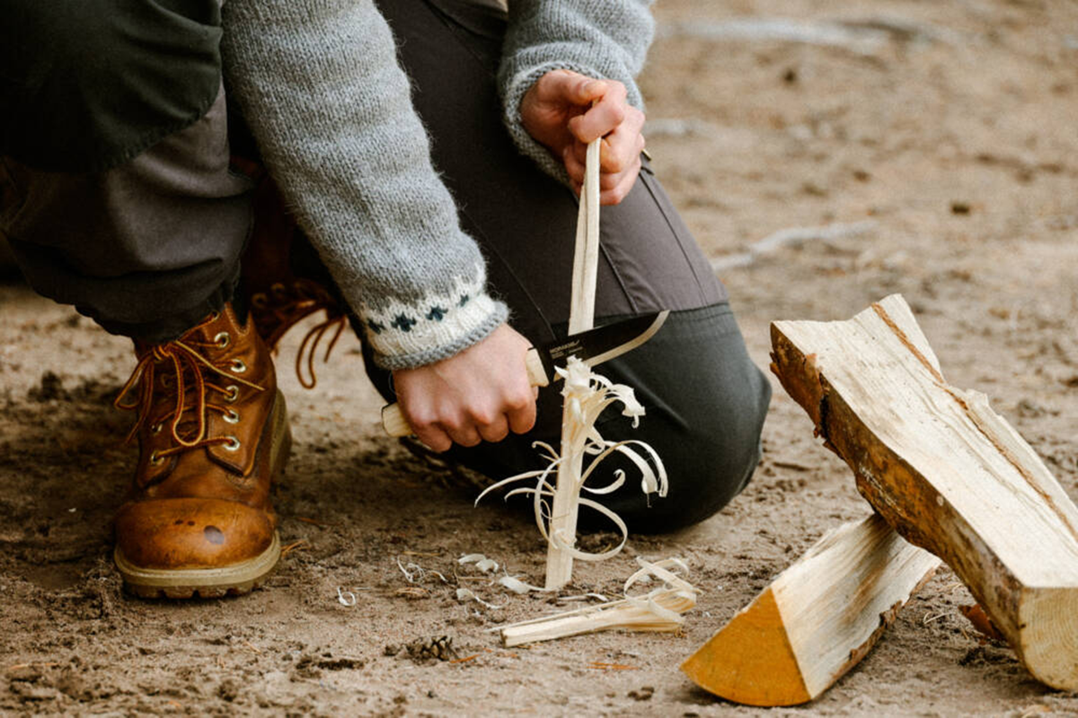Henkilö polvistuu ulkona ja veistää puunpalaa Mora-veitsellä