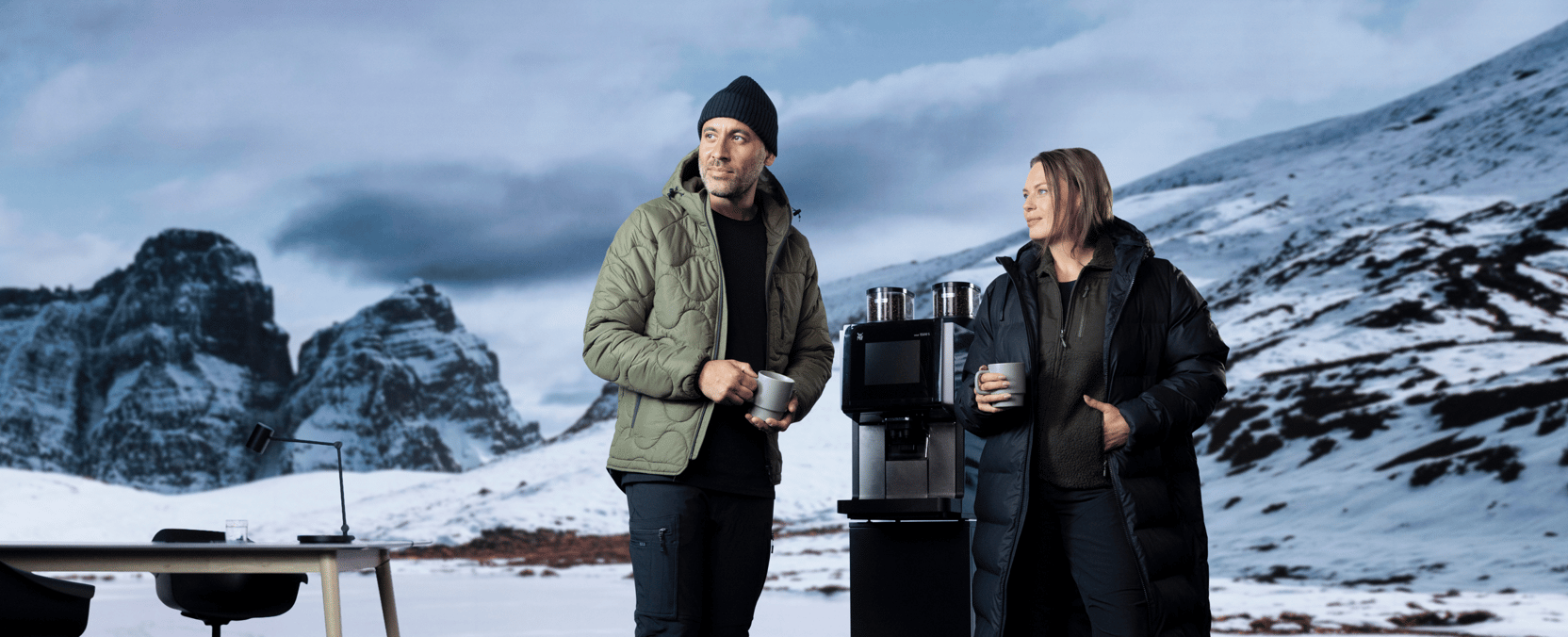 To personer i vinterklær står ved en kaffemaskin utendørs, med snødekte fjell i bakgrunnen og kopper i hendene.