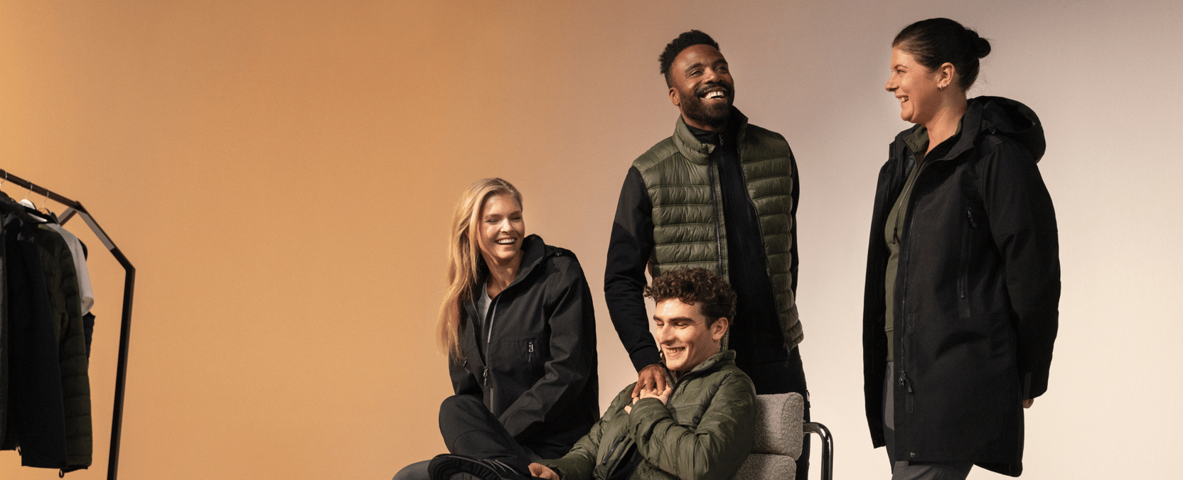 Four people in dark functional clothing laugh together; two are standing and two are sitting in front of a clothing rack.
