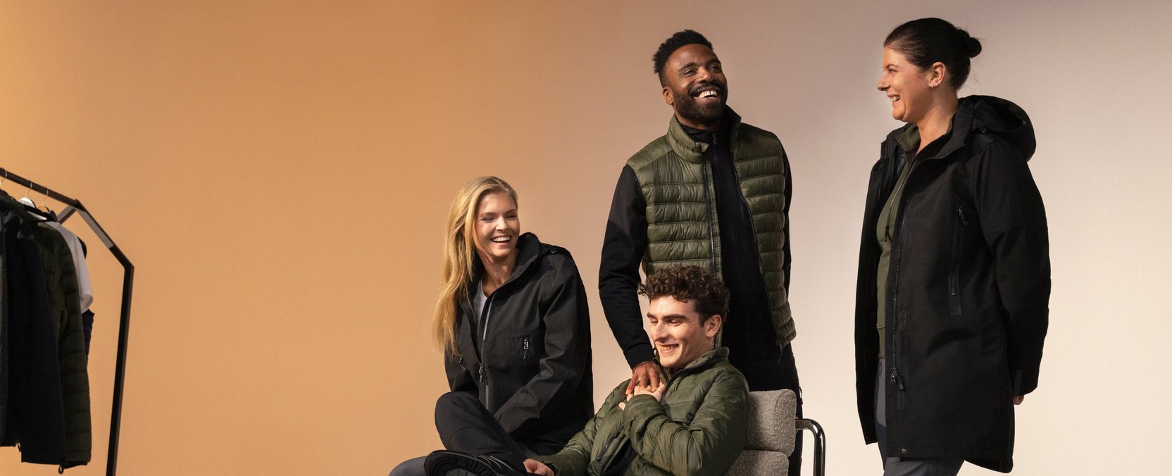 Four people in dark functional clothing laugh together; two are standing and two are sitting in front of a clothing rack.