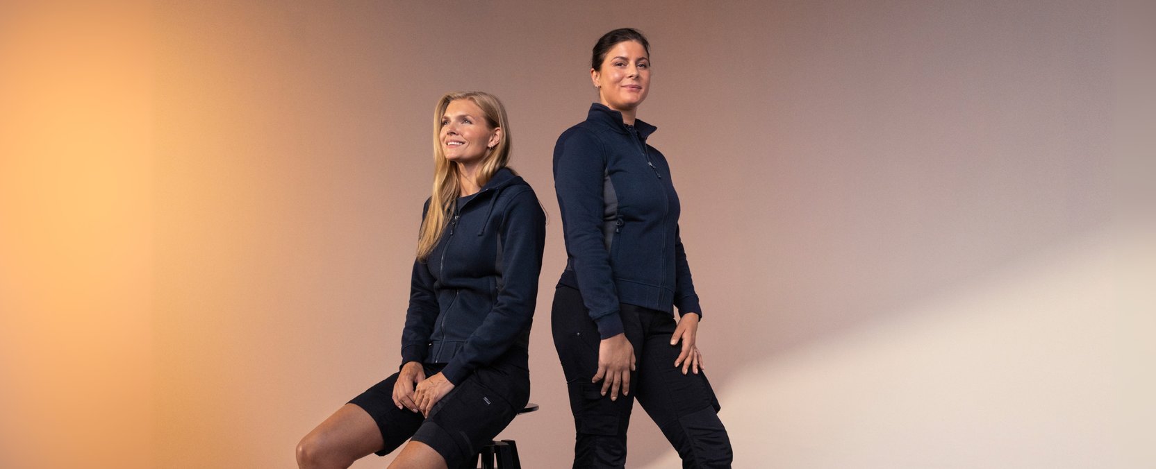 Two women in dark blue performance tops, one sitting and one standing, pose in front of a softly lit background.