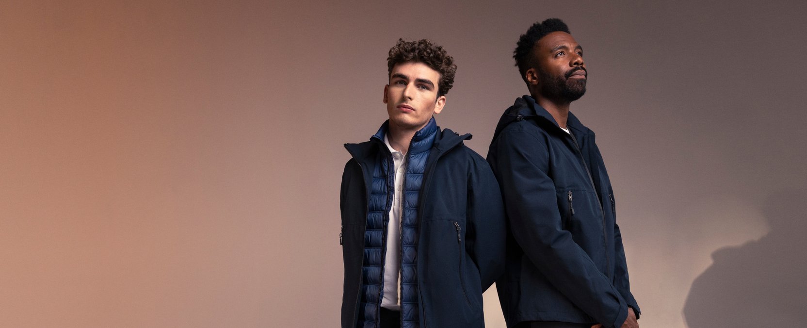 Two men stand back-to-back in dark blue outerwear against a warm, toned background.