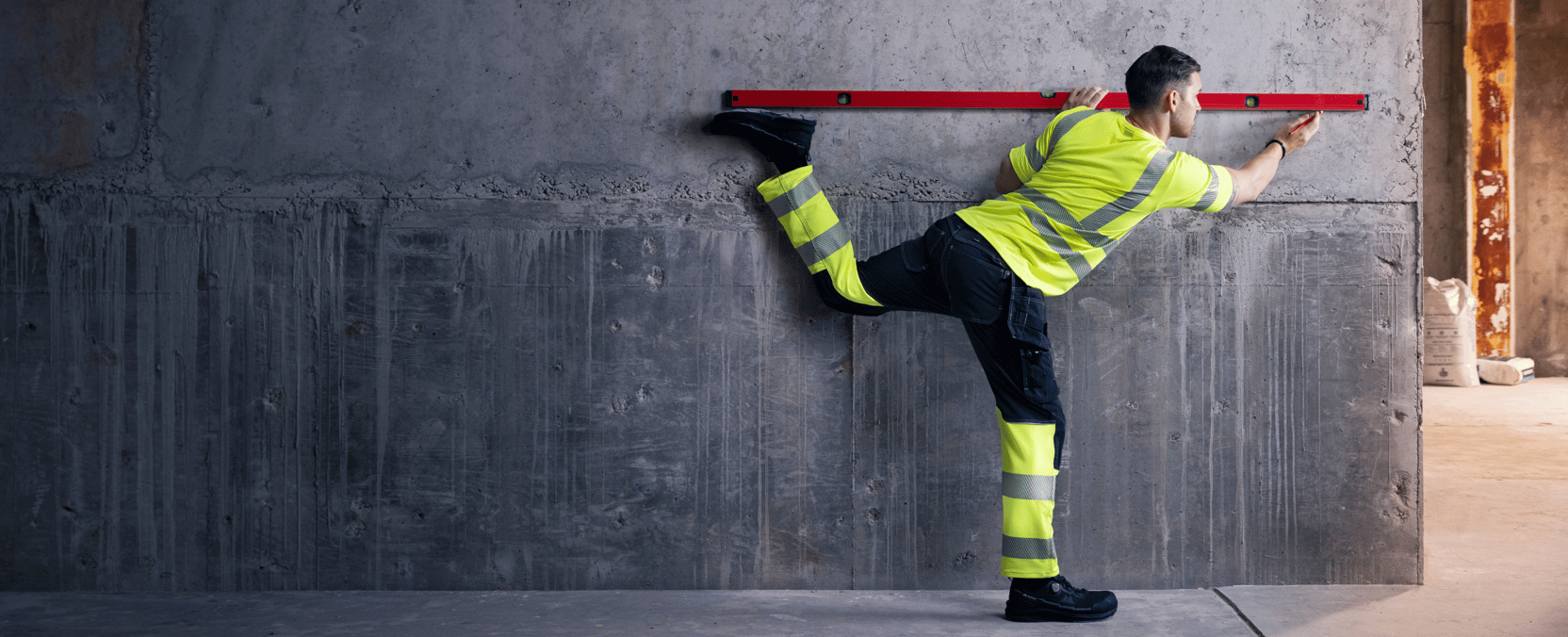 Mand i synlighedstøj står på et ben og balancerer op ad en væg, mens han bruger et vater i øjenhøjde.