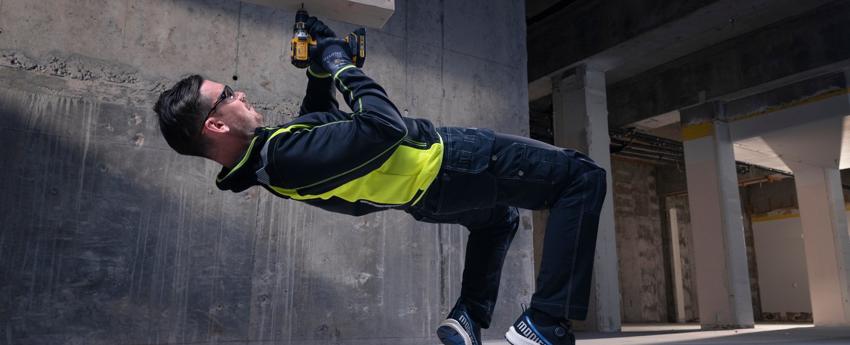 Mand i sorte arbejdsbukser og synlighedstrøje hænger fra loftet med en boremaskine, mens han borer opad.