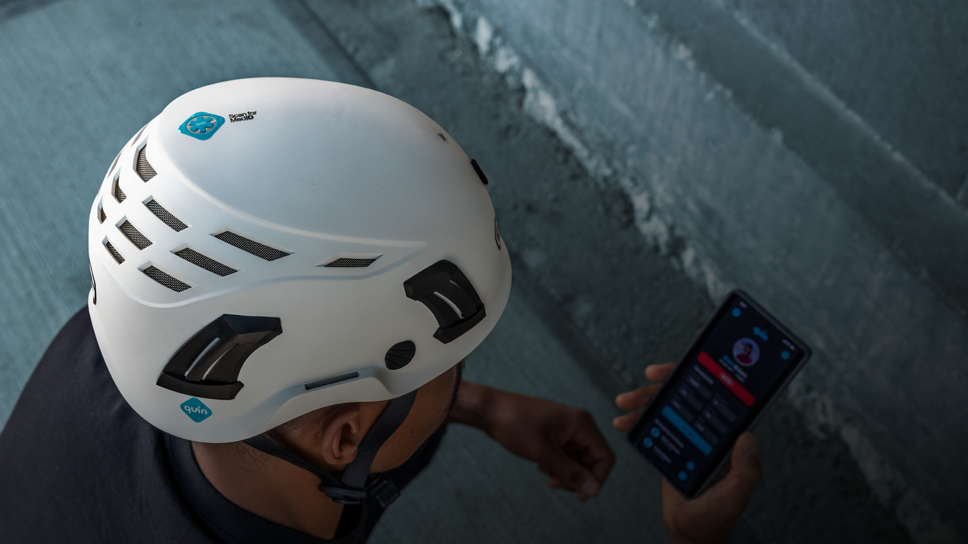 A picture taken from above of a man wearing a white safety helmet with Quin's logos on it. In his hand, he holds a mobile phone with Quin's app