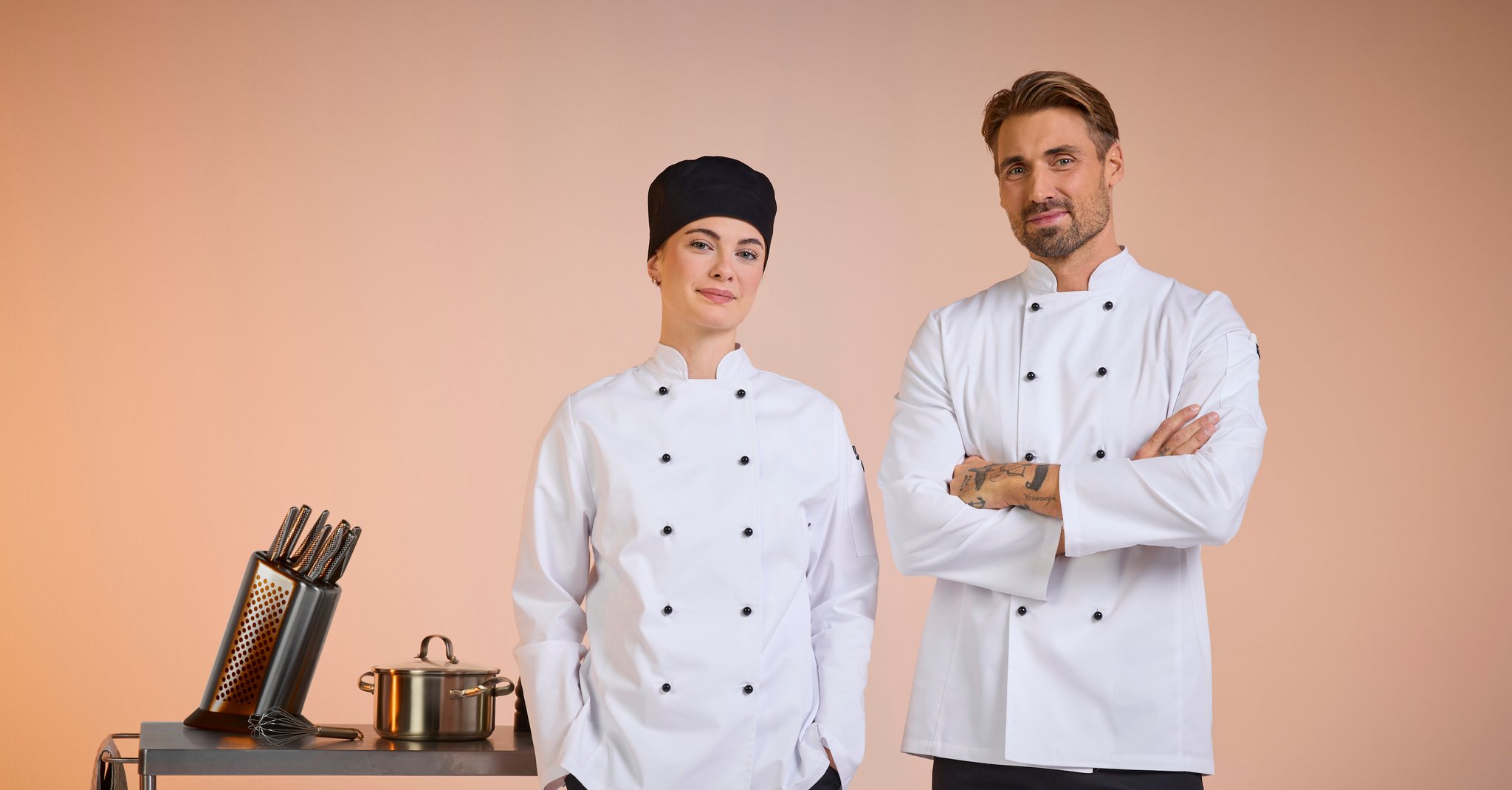 Kvinde og mand i klassiske kokkejakker fra South West, stående i et moderne køkken – professionelt arbejdstøj med stil, komfort og funktion.