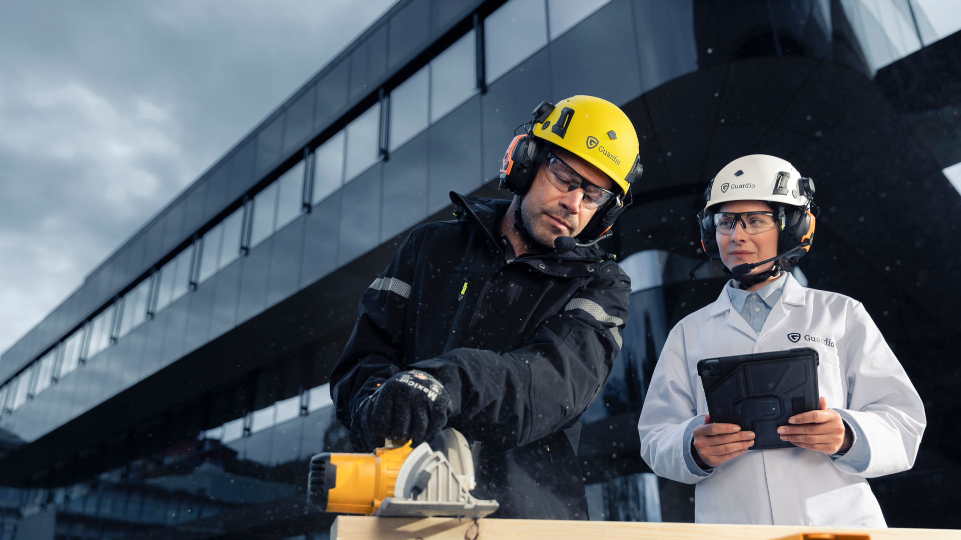 En man i arbetskläder och gul skyddshjälm utför arbete utanför en stor byggnad. Bakom honom står en vetenskapskvinna i vit skyddshjälm och observerar honom