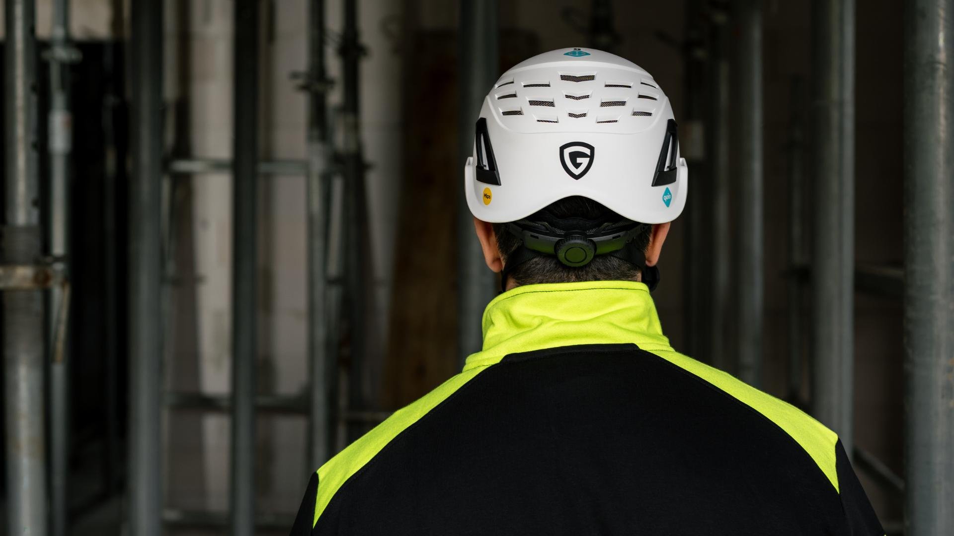 Ein Mann in Arbeitskleidung und gelbem Schutzhelm verrichtet Arbeiten vor einem großen Gebäude. Hinter ihm steht ein Wissenschaftler mit weißem Helm und beobachtet ihn