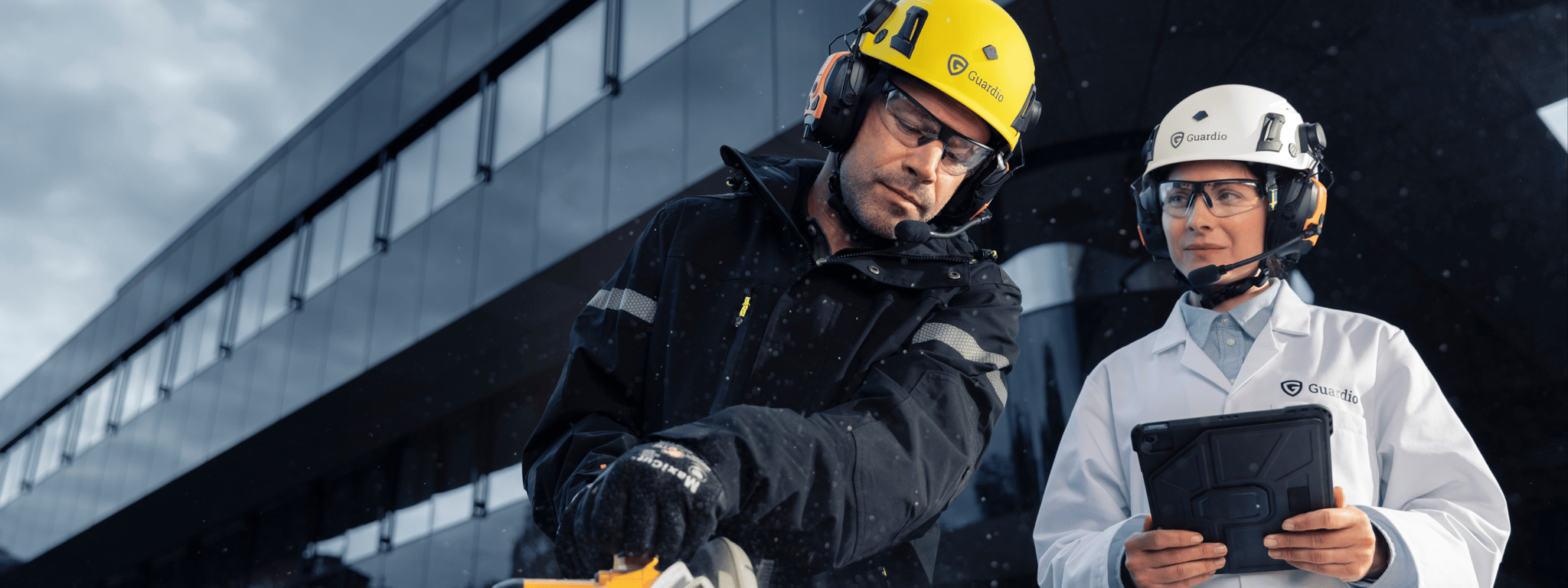 Zwei Personen mit Schutzausrüstung von Guardio – einer im Laborkittel mit Tablet und einer in Arbeitskleidung mit Säge, beide mit Helm, Gehörschutz und Schutzbrille – arbeiten gemeinsam vor einem modernen Gebäude.