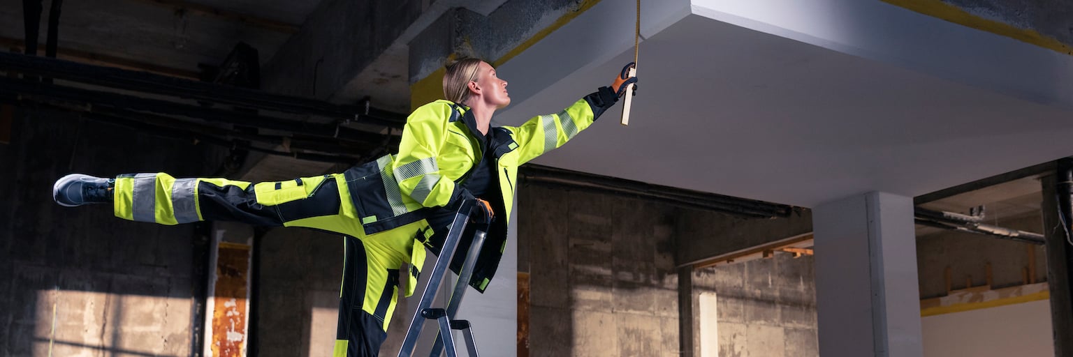 Kvinde i synlighedstøj står højt på en stige med det ene ben strakt bagud og måler loftshøjden med et målebånd.