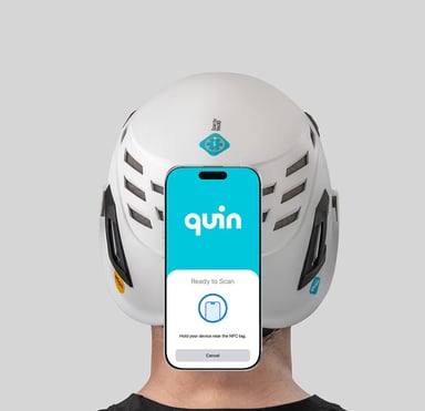 Bakgrund: man klädd i en vit skyddshjälm med Quin-loggan. I förgrunden: en mobiltelefon som visar Quin-appen med texten "Ready to scan".