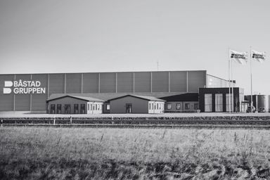 Båstadgruppens hovedkontor og lager i Ängelholm. Moderne industribygg med firmalogo og flagg, fotografert fra utsiden. Bilden kan klikkes på og lenker til mer info om Båstadgruppens historie.