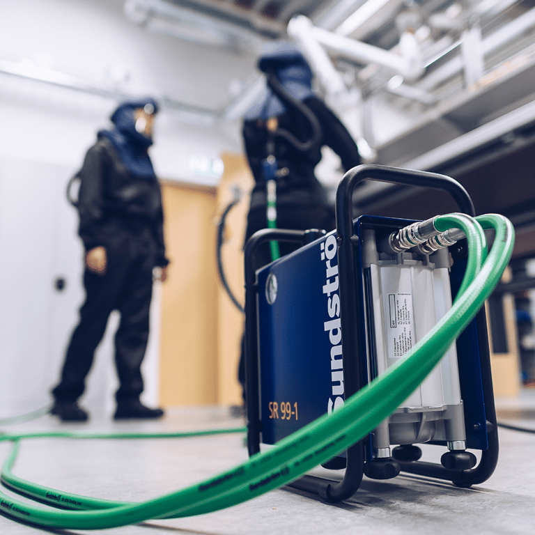 I förgrunden en tryckluftsfilter med slangar kopplade till två arbetare iklädda trycklufts andningsskydd i bakgrunden