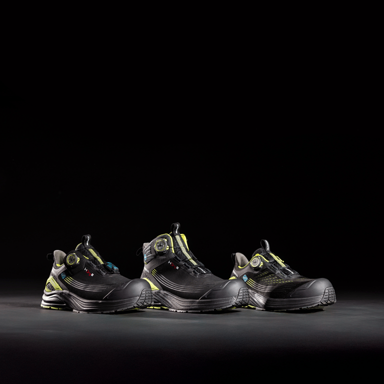 Tre skor ur Monitors ICE Evolution-serie står upplysta mot en mörk, svart bakgrund. Ljuset framhäver skornas design och detaljer, med varje modell placerad bredvid varandra för att visa skillnader i form och utförande.