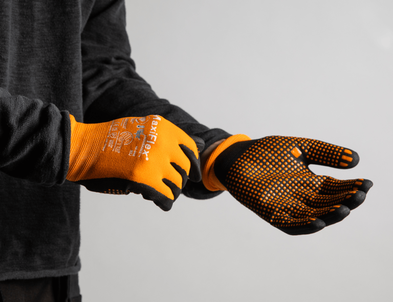 Die Person zieht ein Paar ATG MaxiFlex Endurance Handschuhe an – orangefarbene Handschuhe mit schwarzer Handfläche und Griffverstärkung. Anklickbares Bild mit Link zu ATG-Produkten im Webshop von Båstadgruppen.