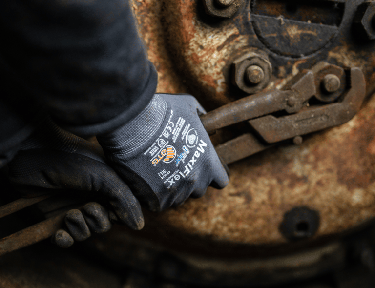 Nahaufnahme von Händen mit ATG MaxiFlex-Handschuhen, die mit rostigen Werkzeugen arbeiten – Langlebigkeit und Präzision in rauen Umgebungen. Anklickbares Bild mit Link zu ATG-Produkten im Webshop von Båstadgruppen.