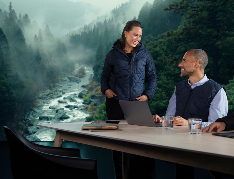 Två kollegor i vadderade Matterhorn-jackor vid ett konferensbord. De ler mot varandra med en forsande älv och granskog i bakgrunden. Klickbar bild, länk till Båstadgruppens hållbarhetsarbete.
