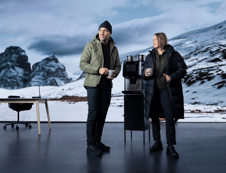 Två personer i vinterjackor står med kaffekoppar vid en kaffemaskin. Bakom dem syns ett snötäckt bergslandskap och ett skrivbord. Klickbar länk till Matterhorn-produkterna.