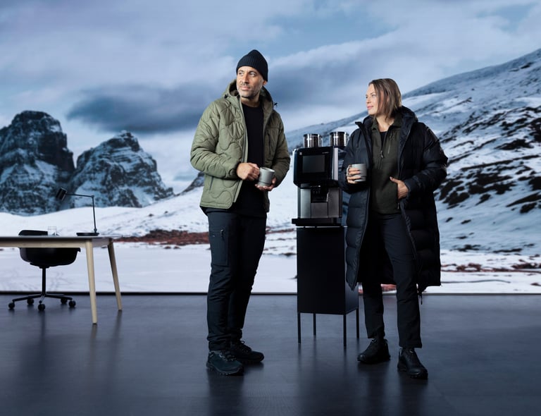 Två personer i vinterjackor står med kaffekoppar vid en kaffemaskin. Bakom dem syns ett snötäckt bergslandskap och ett skrivbord. Klickbar länk till Matterhorn-produkterna.