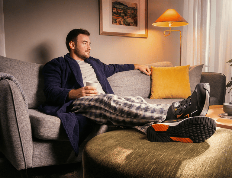 Man i morgonrock sitter avslappnat i soffa med kopp kaffe, visar undersidan på Monitor-skyddsskor med orange detaljer – klickbar länk