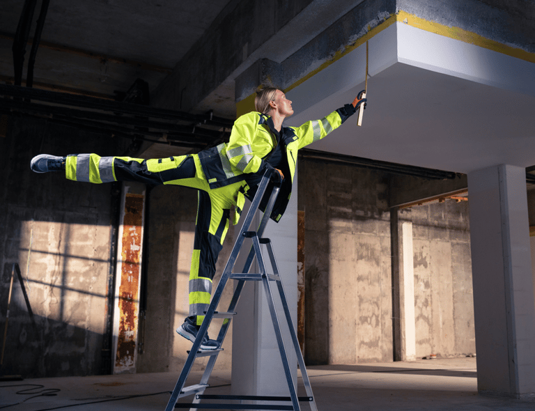Nainen varoitusvaatteissa seisoo tikkailla toinen jalka ojennettuna taakse balettiasennossa mitatessaan kattokorkeutta – Top Sweden työvaatteet taipuvat mukana.