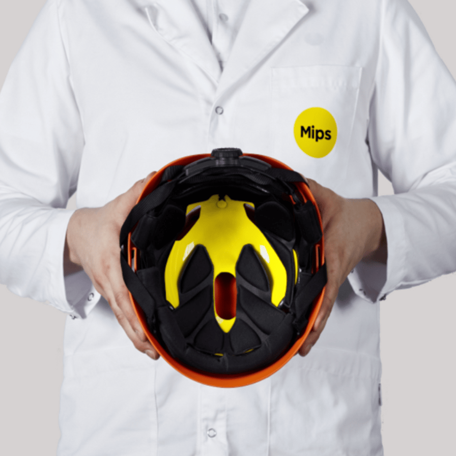 A person in a white lab coat with the Mips logo is holding a safety helmet with a yellow Mips system inside