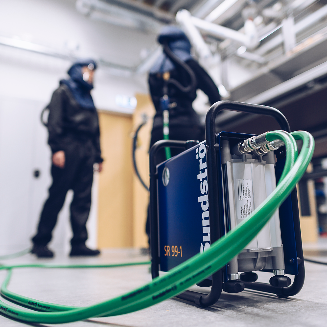 Im Vordergrund ein Druckluftfilter mit Schläuchen, die mit zwei Arbeitern verbunden sind, die im Hintergrund Druckluft-Atemschutzmasken tragen