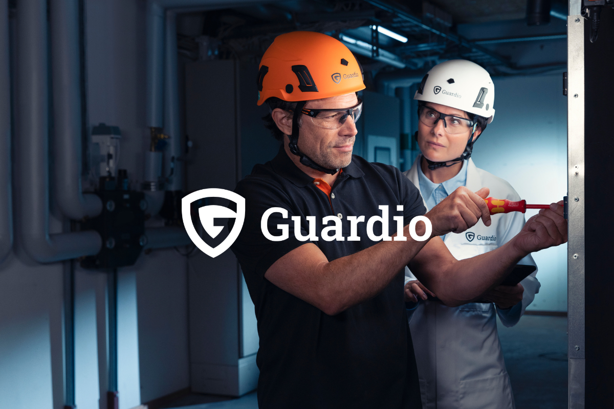 Two people with safety helmets working together in a technical environment, one is screwing into a metal frame. Clickable link to Guardio products
