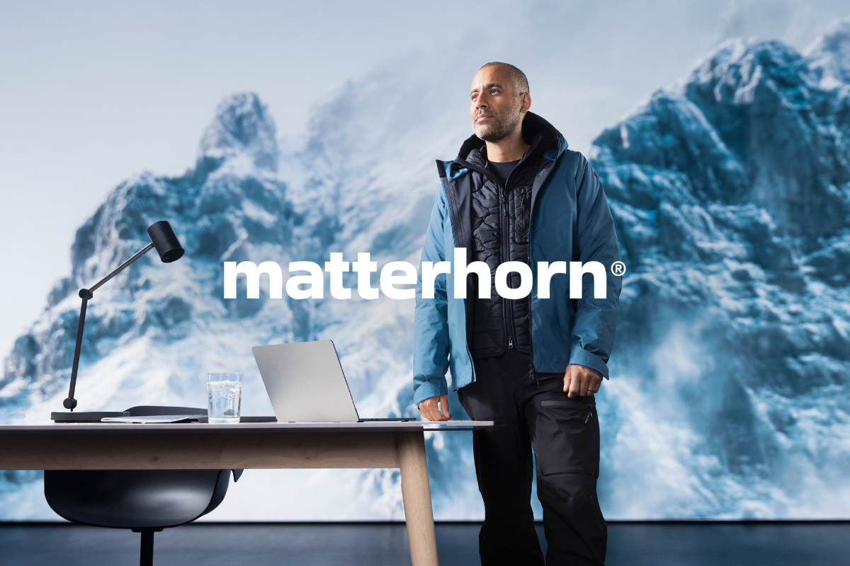 Man in functional clothing standing by a desk with a laptop, snow-covered mountains in the background. Clickable link to Matterhorn products