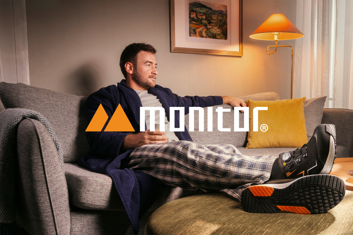 Man in bathrobe and pajamas sitting on a couch with a coffee cup, showing a pair of black safety shoes. Clickable link to Monitor products