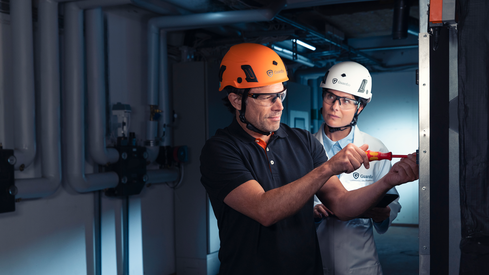 En mand i arbejdstøj og orange sikkerhedshjelm udfører arbejde. Bag ham står en kvindelig videnskabsmand og observerer ham.