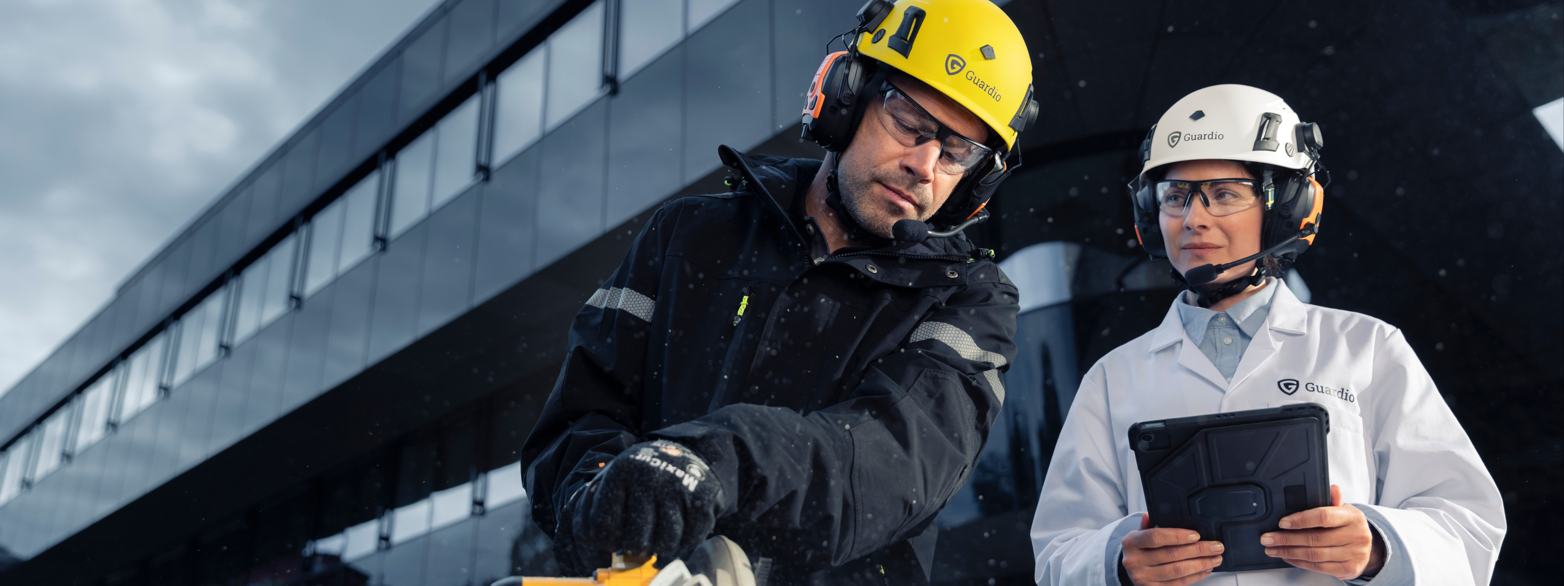 Two people wearing Guardio safety gear – one in a lab coat with a tablet and one in workwear with a saw. Both wear helmets, hearing protection and safety glasses – working together outside a modern building.