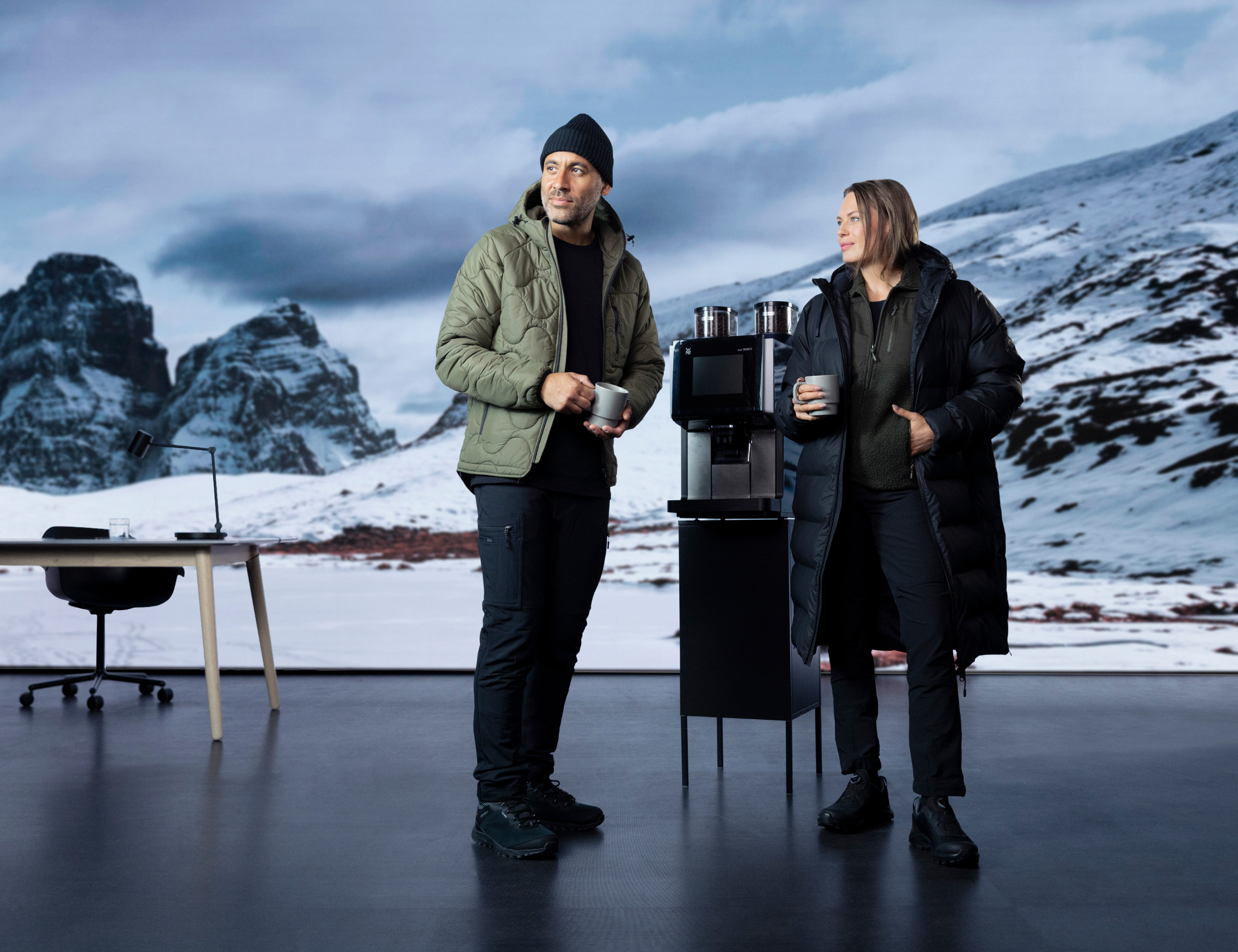 To personer i vinterjakker står med kaffekopper ved en kaffemaskine. Bag dem ses et snedækket bjerglandskab og et skrivebord. Klikbar link til Matterhorn-produkterne.