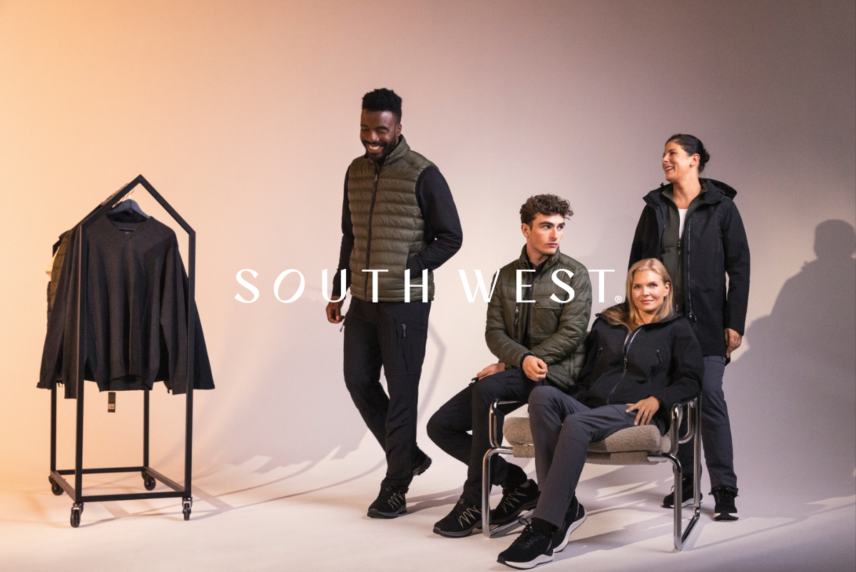 Four people in relaxed workwear posing in a studio, with a clothing rack in the background. Clickable link to South West products