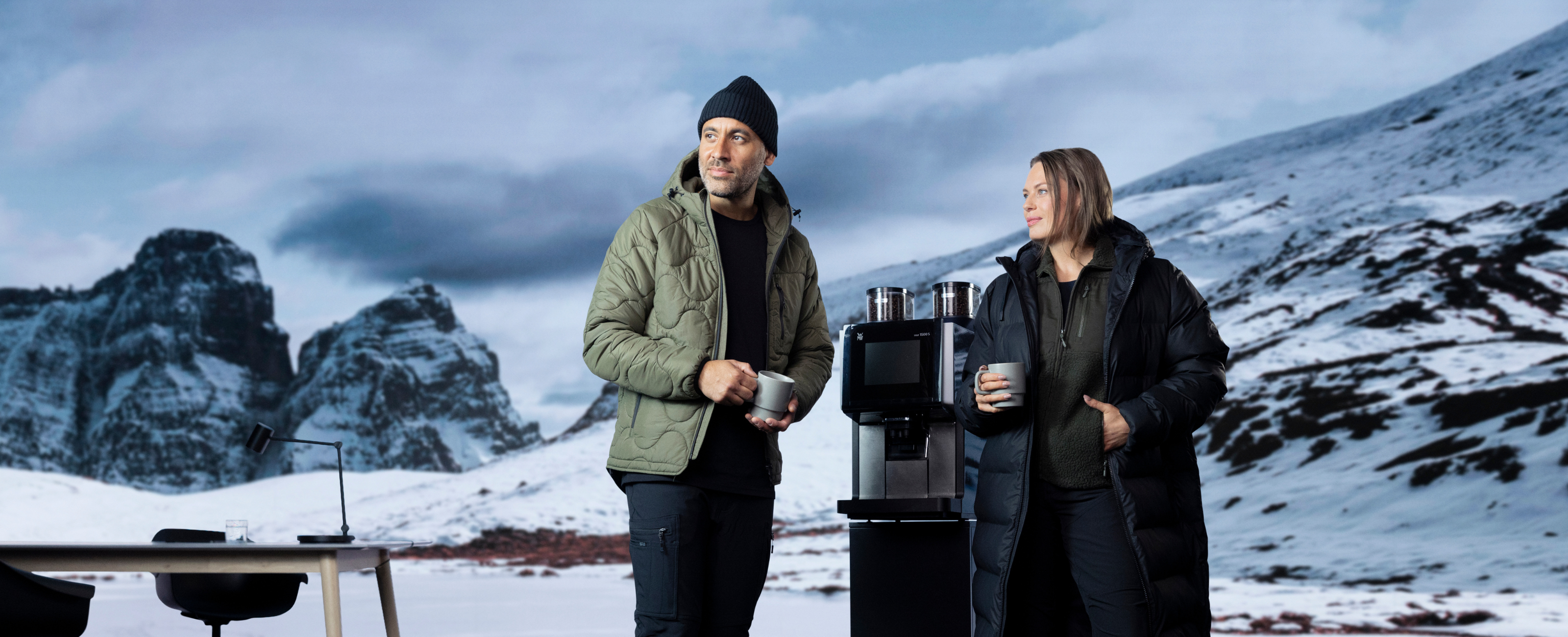 To personer i vinterklær står ved en kaffemaskin utendørs, med snødekte fjell i bakgrunnen og kopper i hendene.